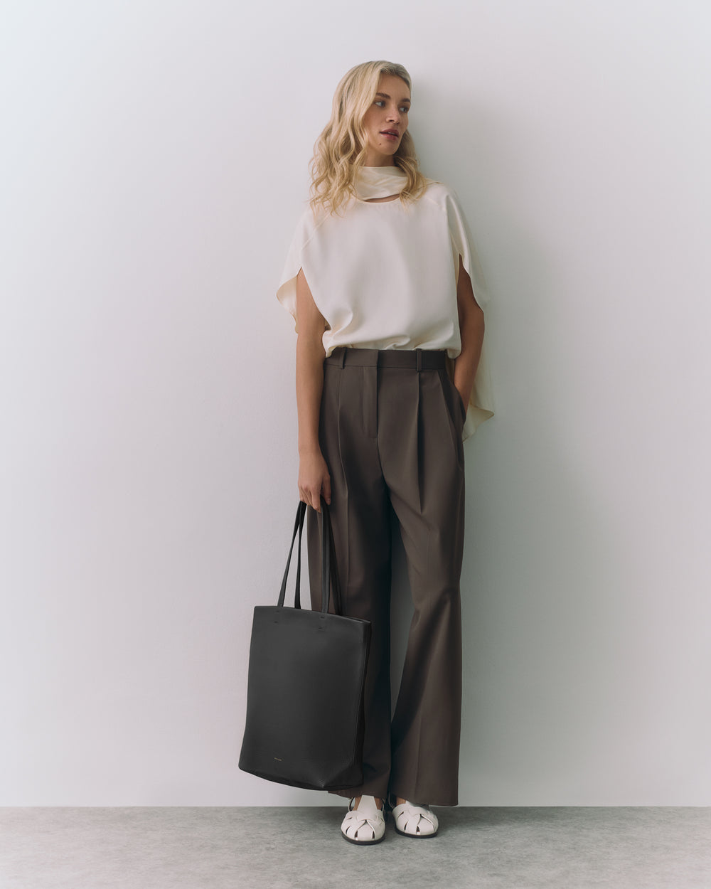 Person wearing a top and trousers holding a tote bag, standing against a plain background.