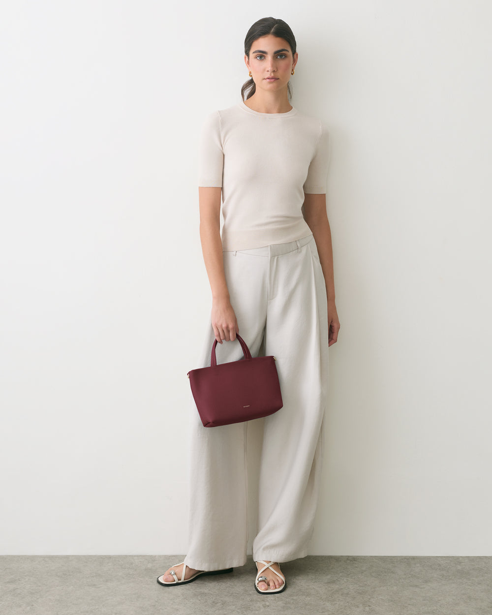 Person stands against wall holding a handbag wearing a short-sleeve shirt and wide-leg pants.