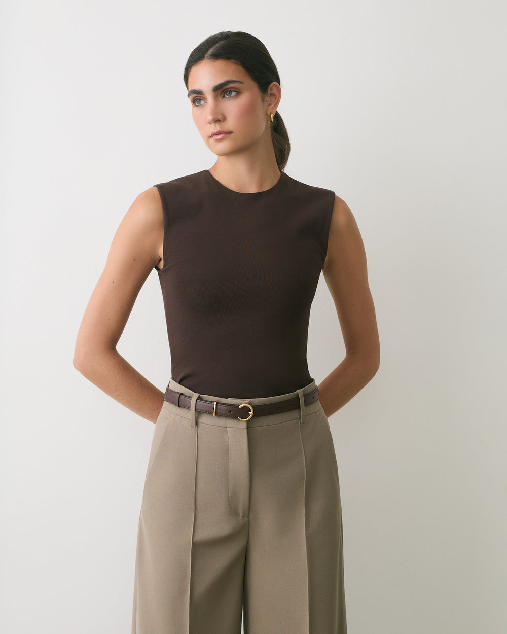Person wearing sleeveless top and pants with belt standing against plain backdrop.