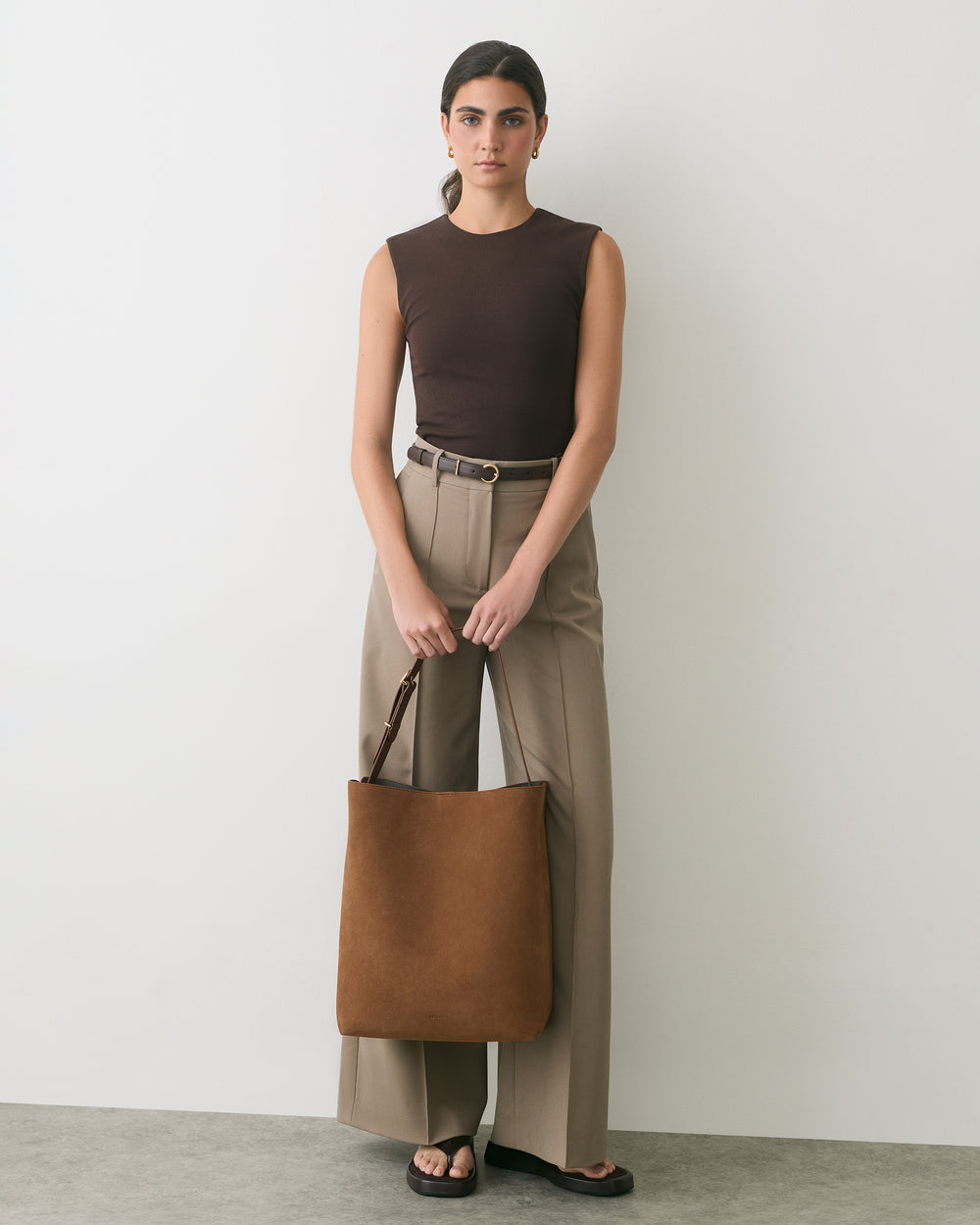 Person standing with tote bag, wearing sleeveless top, pants, and sandals.