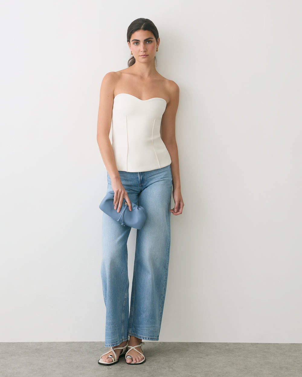 Person in a strapless top and wide-leg jeans holding a small clutch standing against a wall.