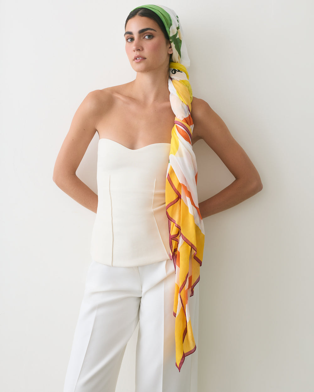 Person wearing a strapless top, pants, and a headscarf standing against a wall.