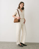 Person standing with shoulder bag, wearing a long-sleeve top and pants, looking slightly to the side.