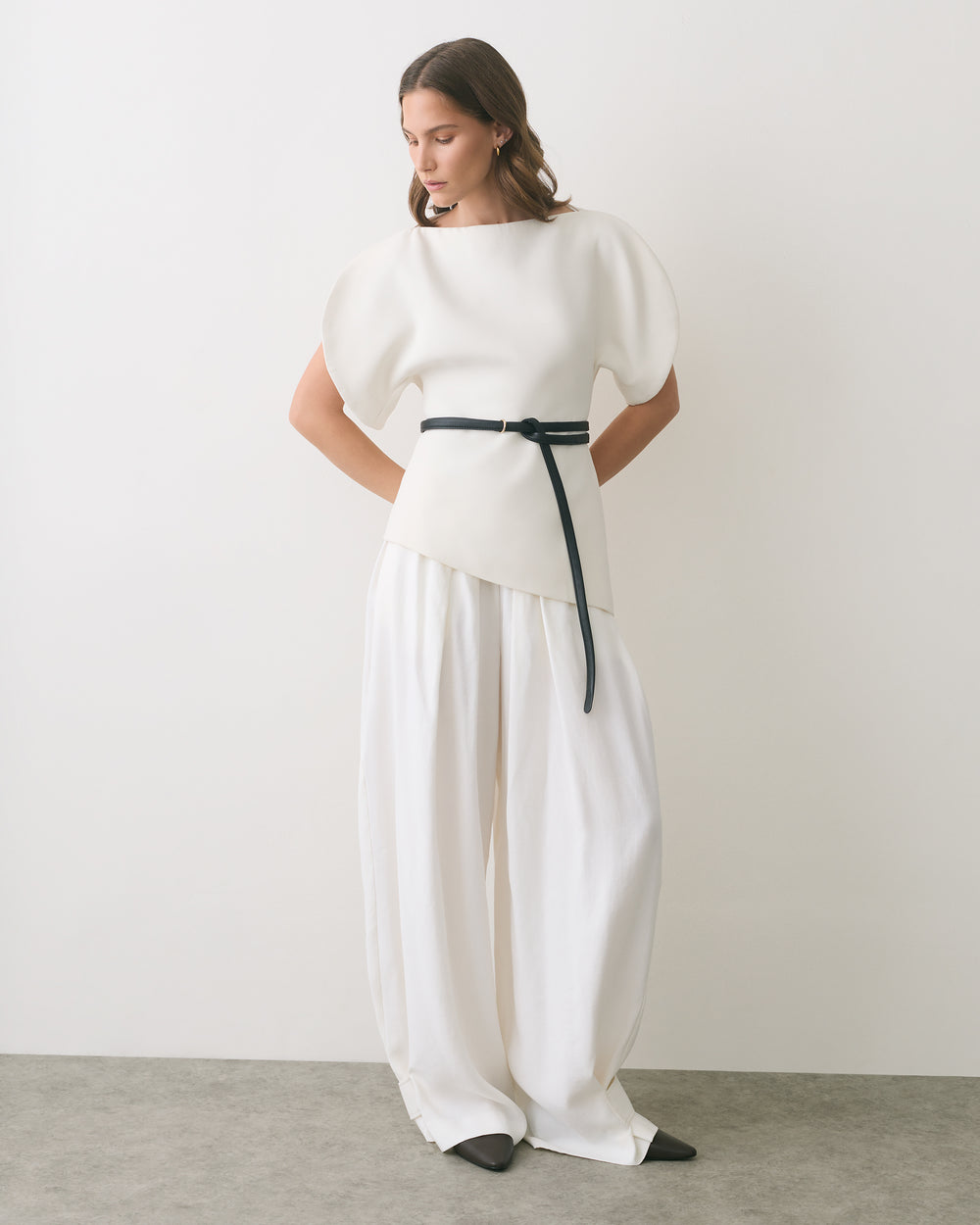 Model wearing loose pants and a top with a belt standing in a neutral setting.