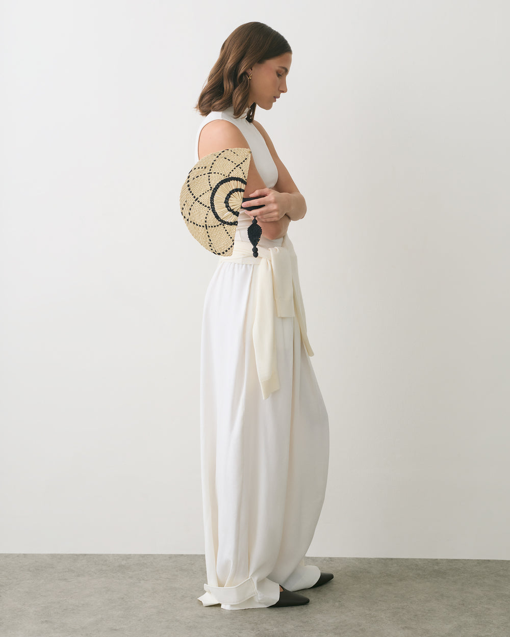 Woman in long outfit holding a round woven bag, standing sideways against plain backdrop.