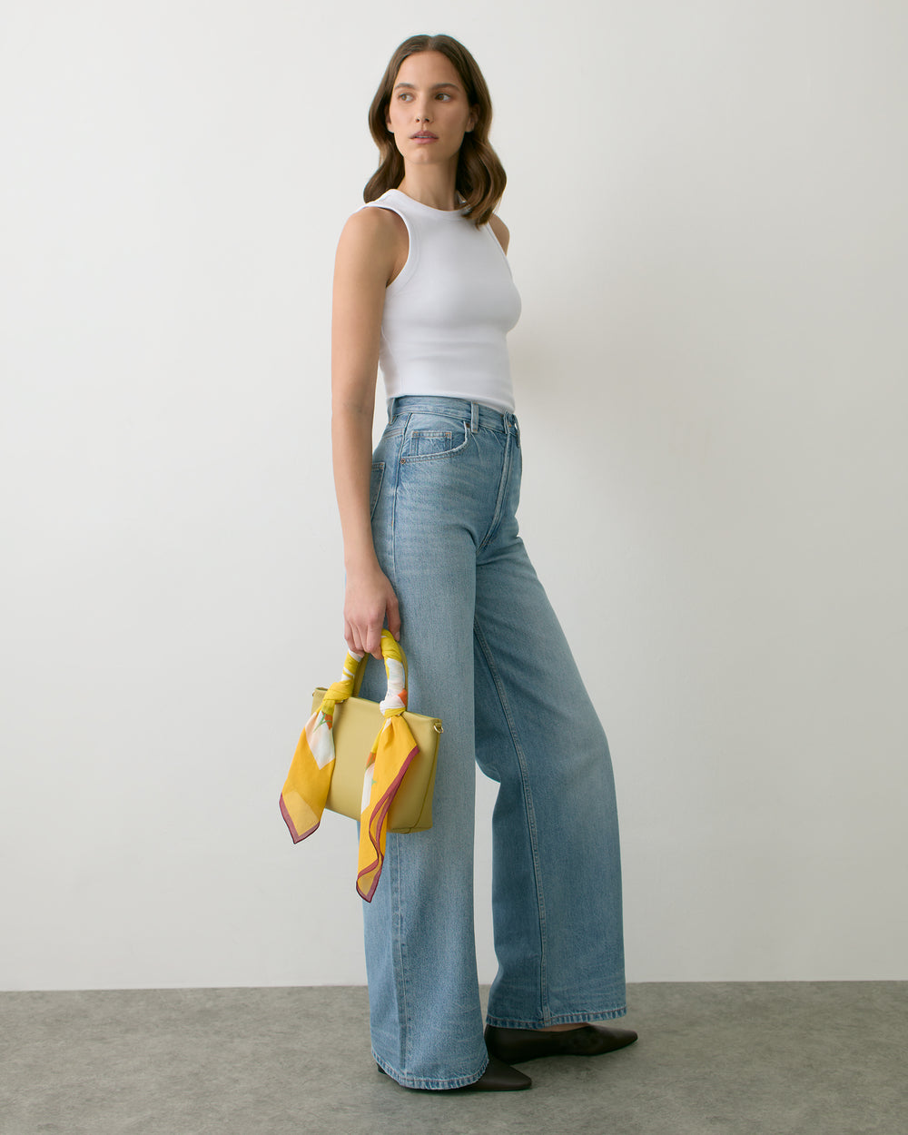 Person in a sleeveless top and wide-leg jeans holding a handbag with scarf detailing.