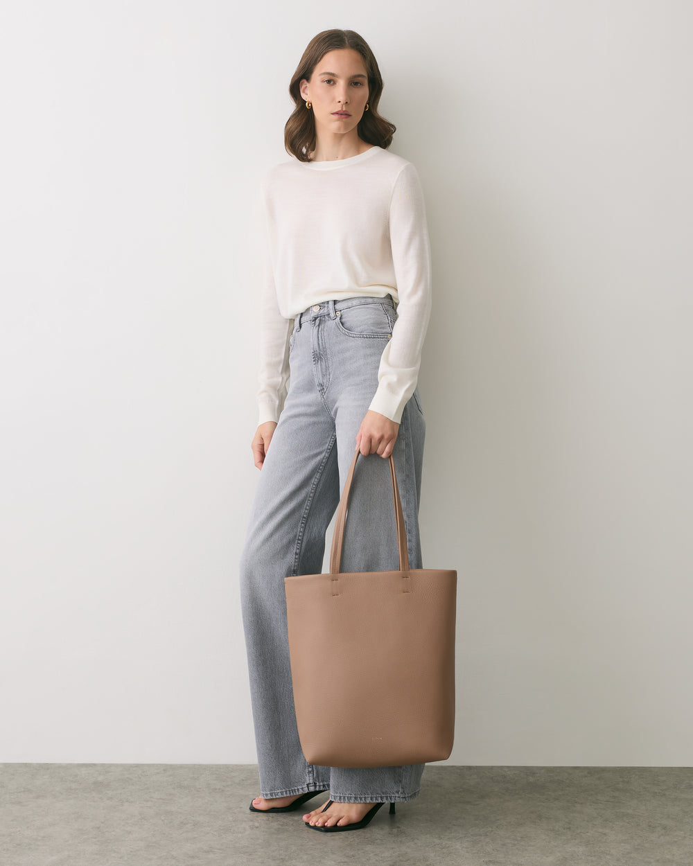 Person holding a tote bag, wearing a long-sleeve top and jeans, standing indoors.