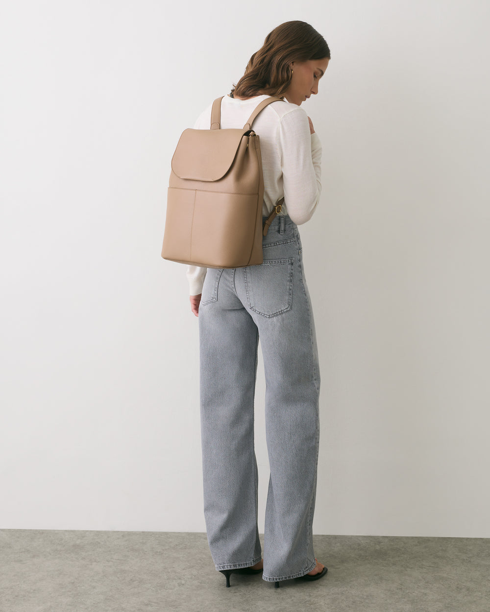 Person wearing jeans and carrying a backpack, facing a white wall.