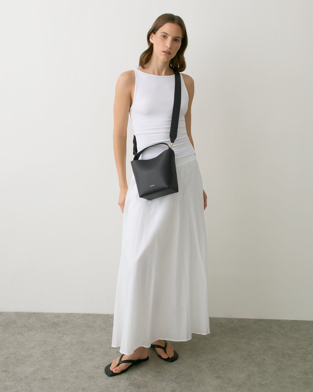 Person wearing a long skirt, tank top, sandals, and crossbody bag standing against a plain wall.