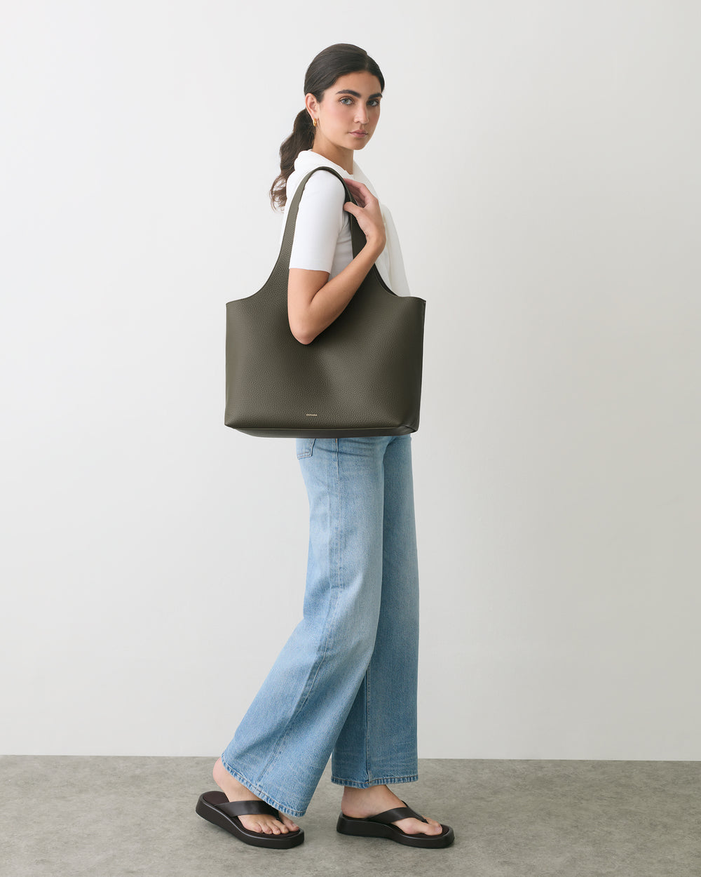 Person carrying a large shoulder bag, wearing jeans and sandals in a minimalist setting.