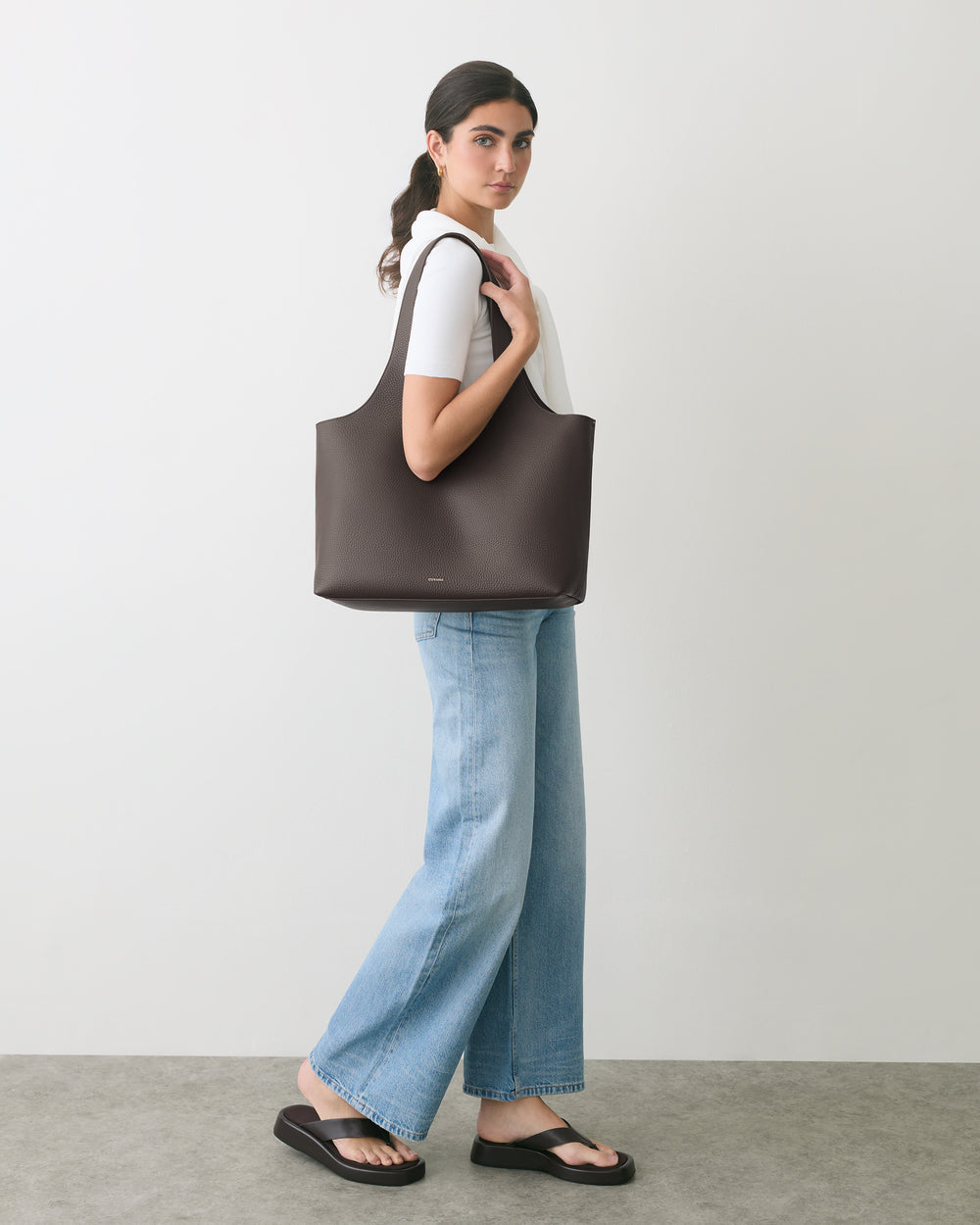 Person standing with tote bag over shoulder, wearing jeans and a t-shirt, looking at camera.