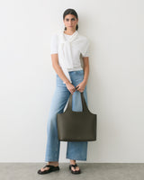 Person with a large tote bag, wearing a shirt and jeans, stands against a plain background.