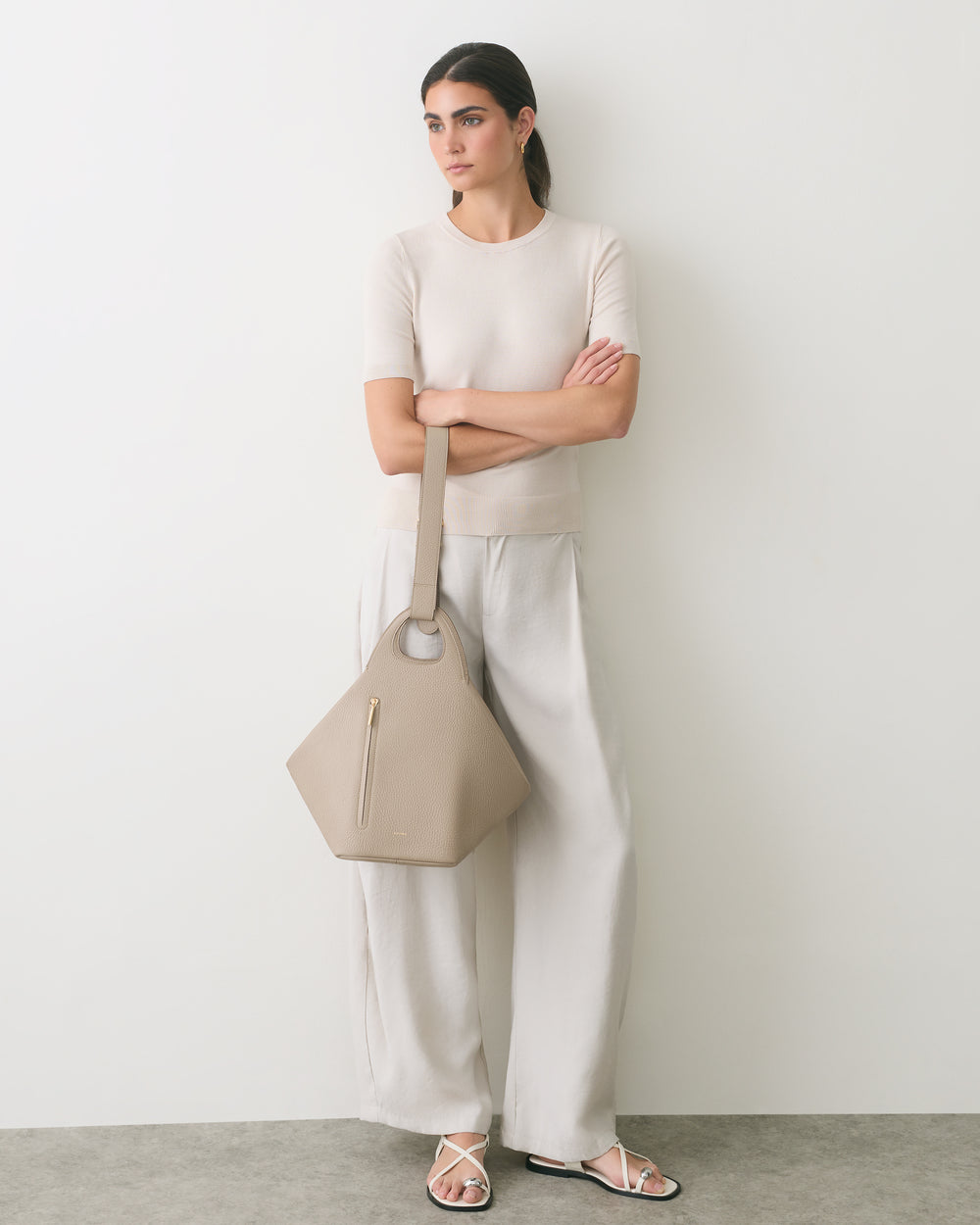 Person in a simple outfit holding a large handbag, standing against a plain wall.