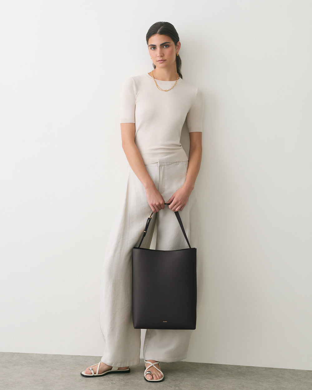 Person standing against a plain background holding a shoulder bag in front of them.