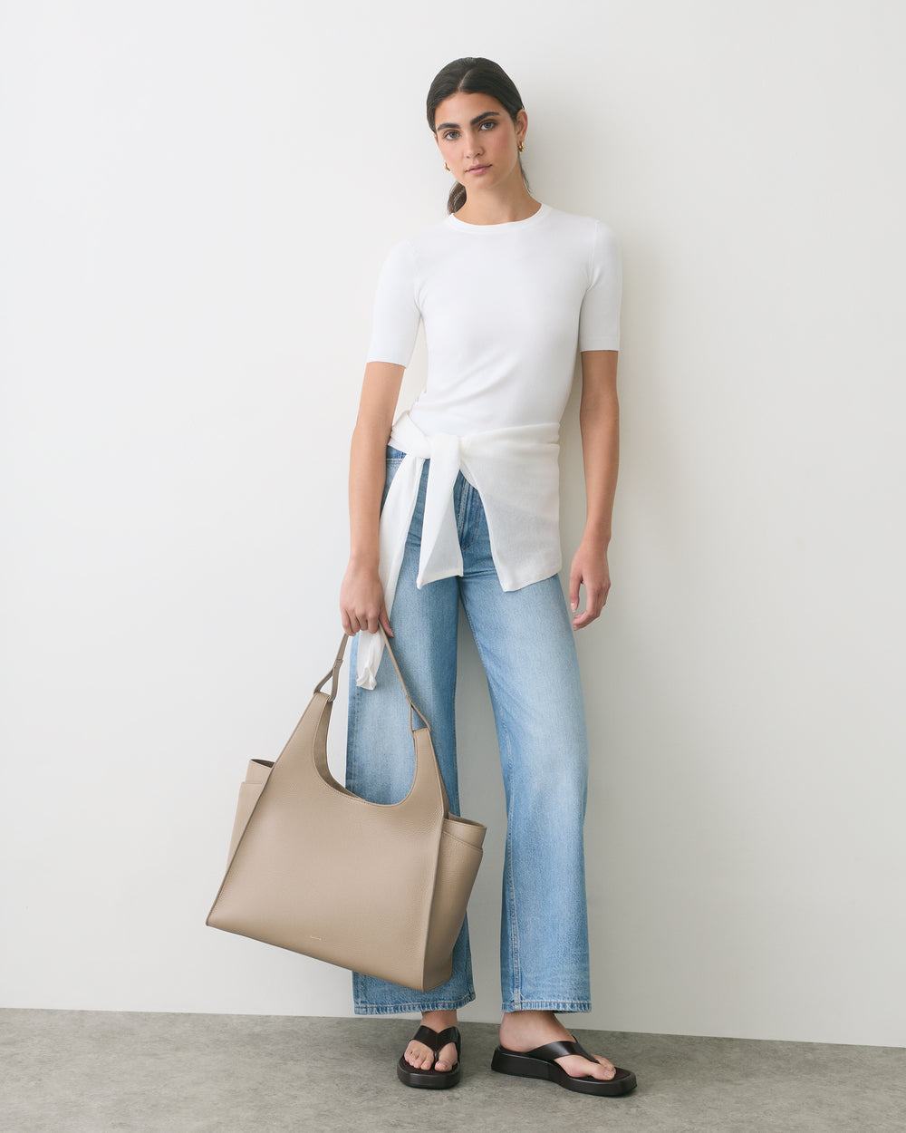Person wearing casual outfit holding a large handbag, standing against a plain wall.