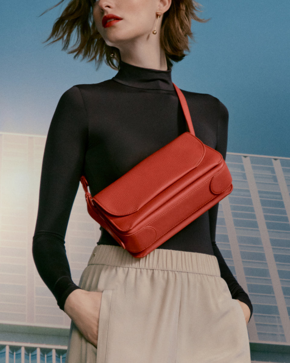 Woman with handbag standing against a sky and building backdrop.