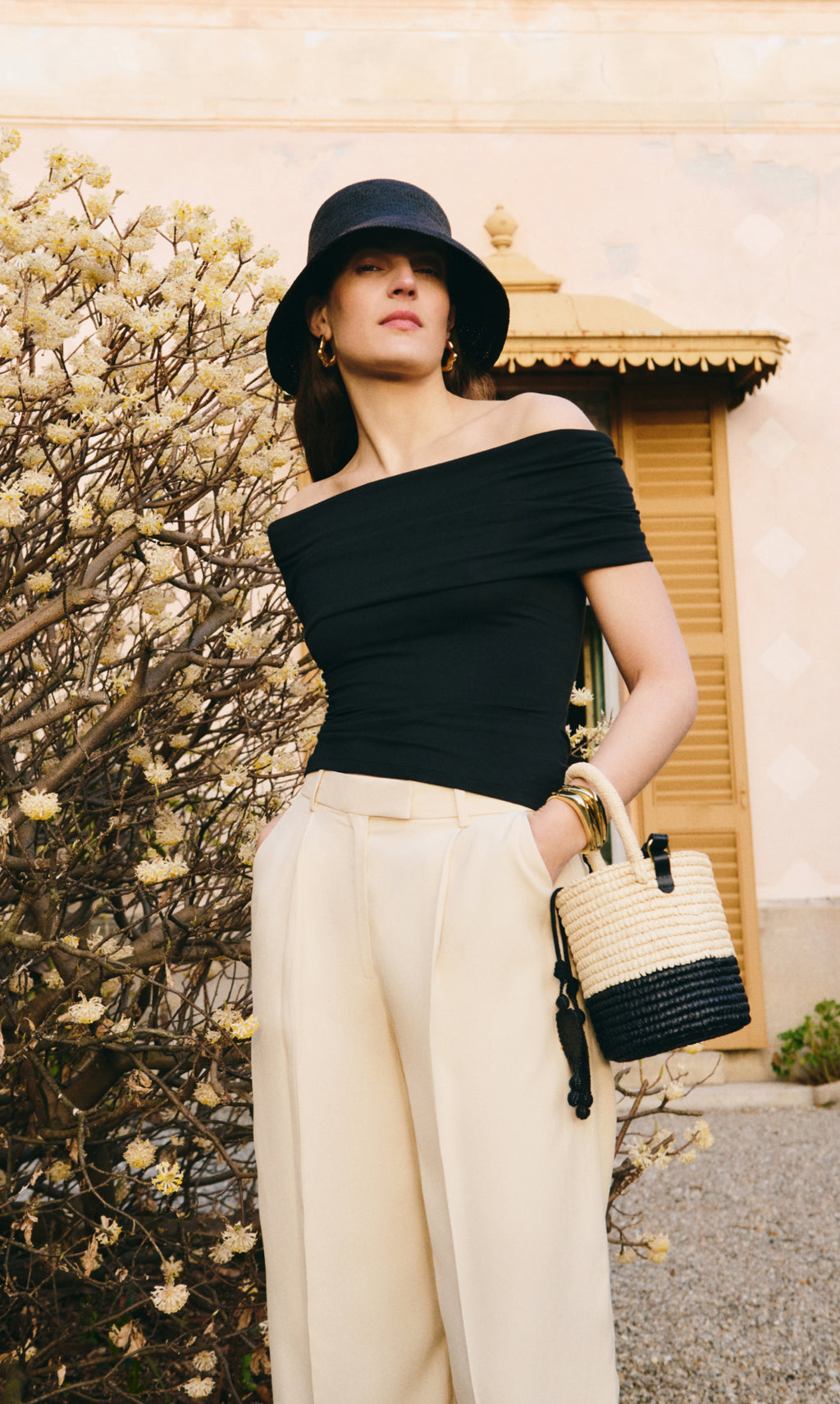 Woman in an off-shoulder top and pants, wearing a hat, holding a woven handbag.