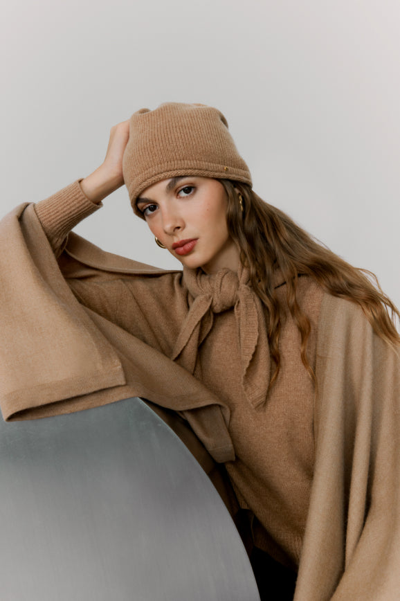 Woman in a beanie leaning on a chair, looking at the camera.