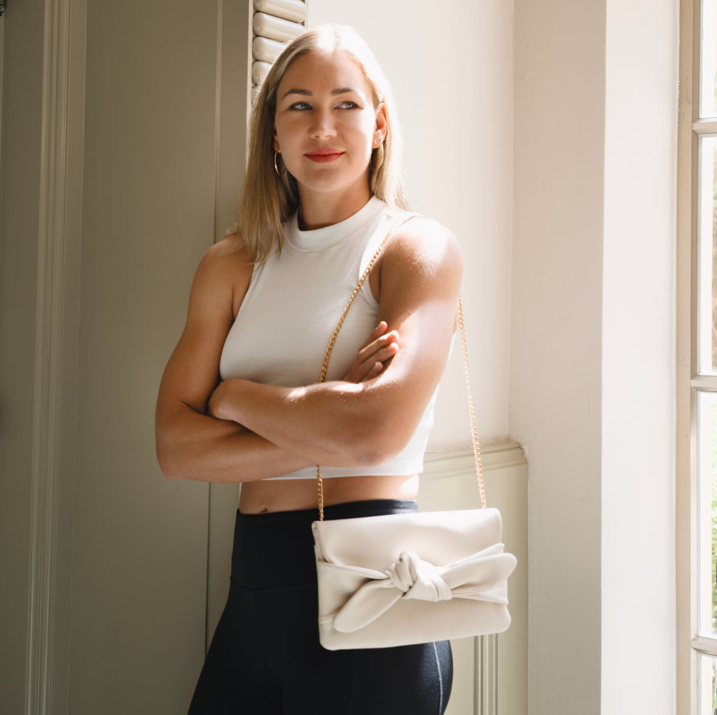 Woman standing by window with arms crossed, holding a purse.