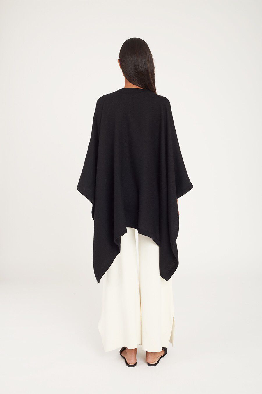 Woman from behind wearing a cape and wide-leg pants, standing in studio.