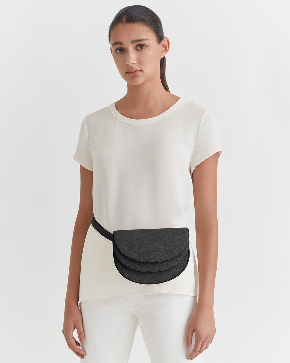 Woman standing in a simple top and pants with a small bag across her shoulder.