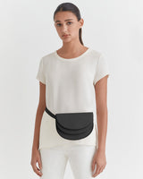 Woman standing in a simple top and pants with a small bag across her shoulder.