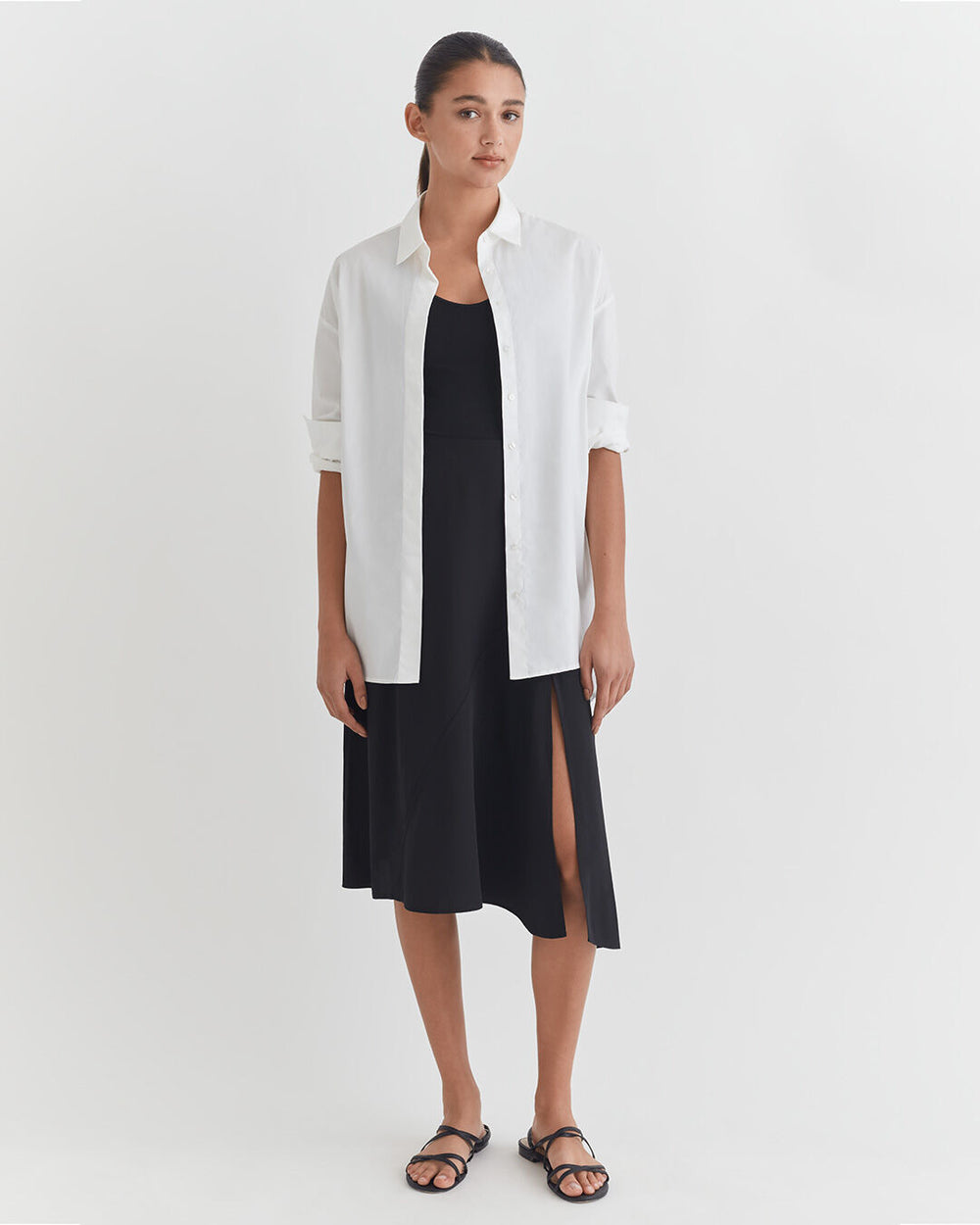 Woman standing in a dress with a button-up shirt over it and sandals.