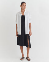 Woman standing in a dress with a button-up shirt over it and sandals.