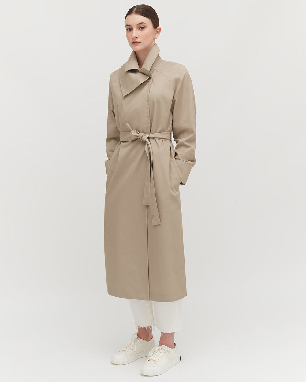 Woman standing in a long coat with a belt, paired with trousers and sneakers.