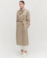 Woman standing in a long coat with a belt, paired with trousers and sneakers.