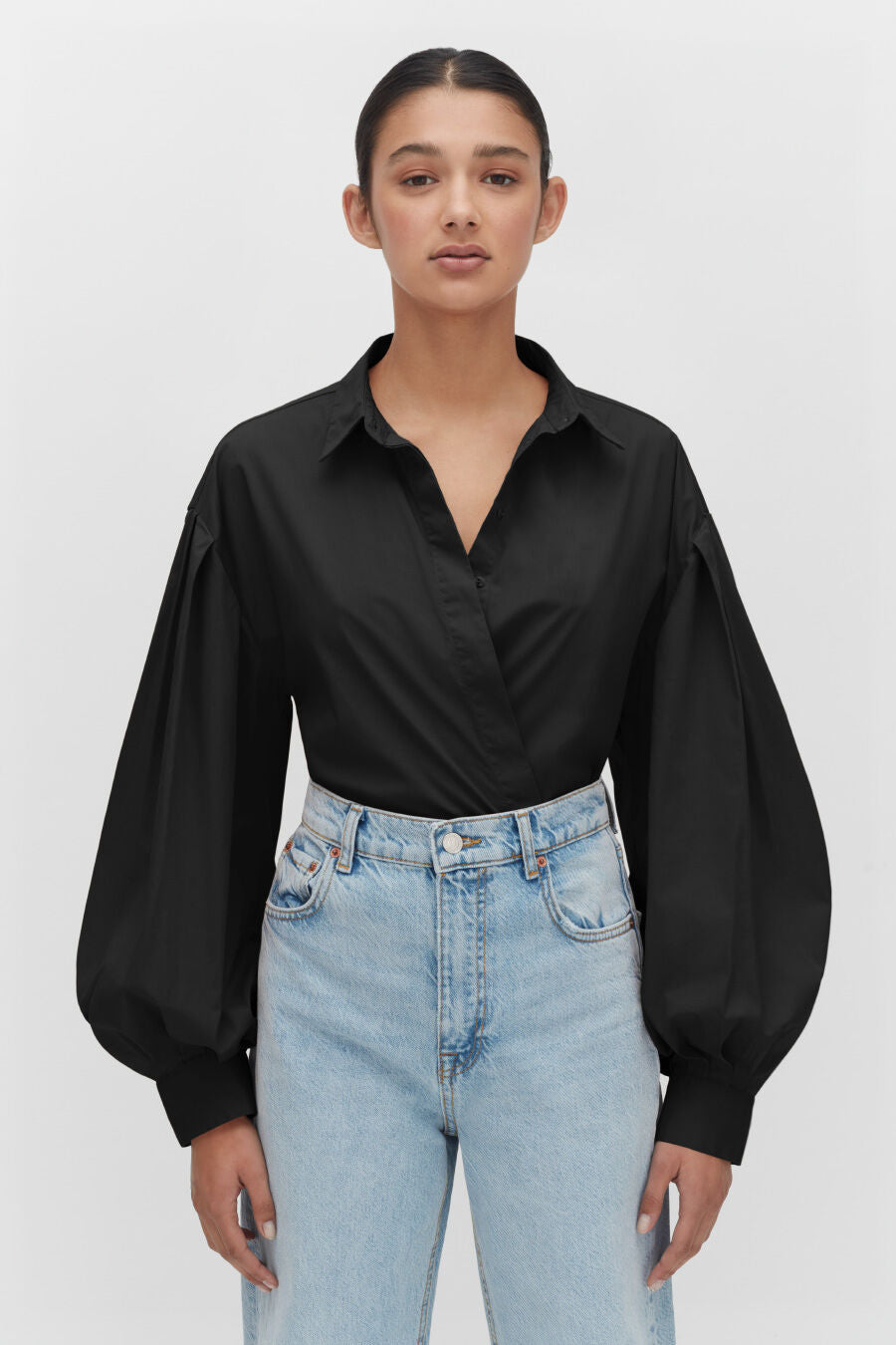 Woman with hands on hips wearing a blouse and jeans, standing against a plain background.
