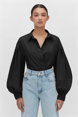 Woman with hands on hips wearing a blouse and jeans, standing against a plain background.