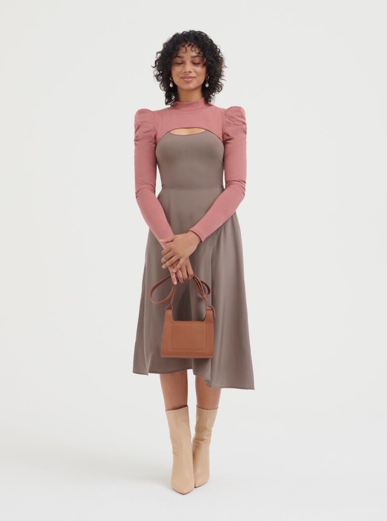 Woman standing with a handbag, wearing a long-sleeve dress and boots.