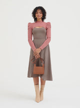 Woman standing with a handbag, wearing a long-sleeve dress and boots.